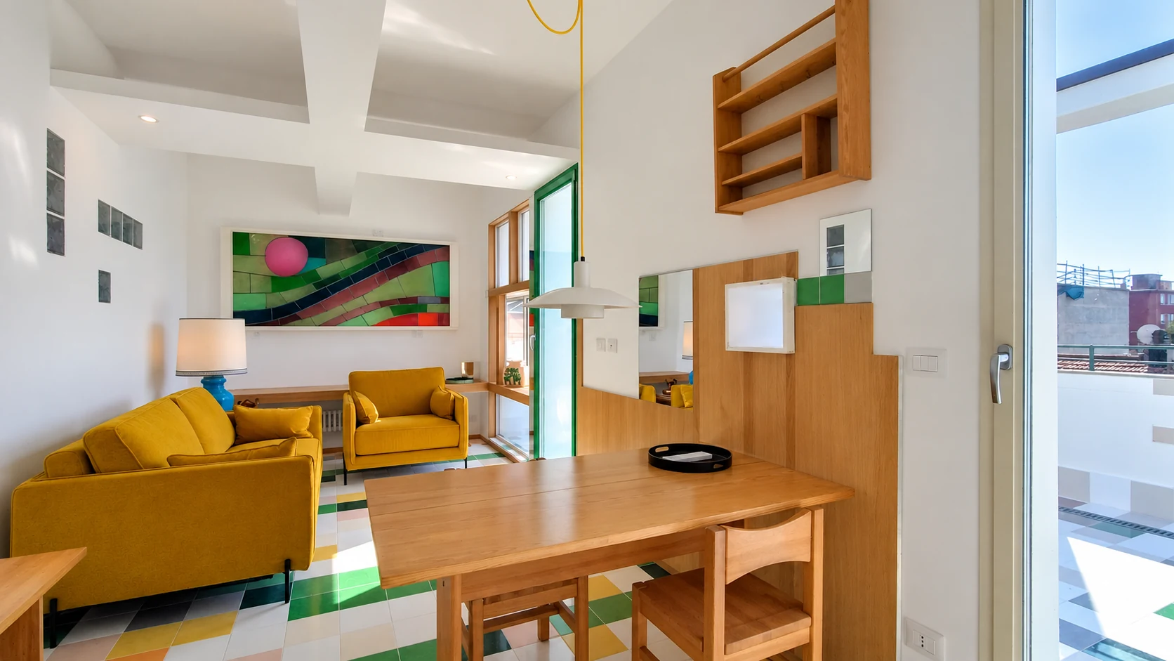 Zona giorno dell’attico con tavolo in legno, sedute gialle e pavimento multicolore, illuminata dalle vetrate sulla terrazza.