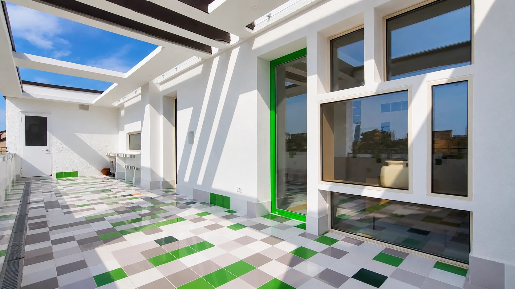 Terrazza con aperture di diverse dimensioni e porta verde, attraversata dalle ombre geometriche della pergola.