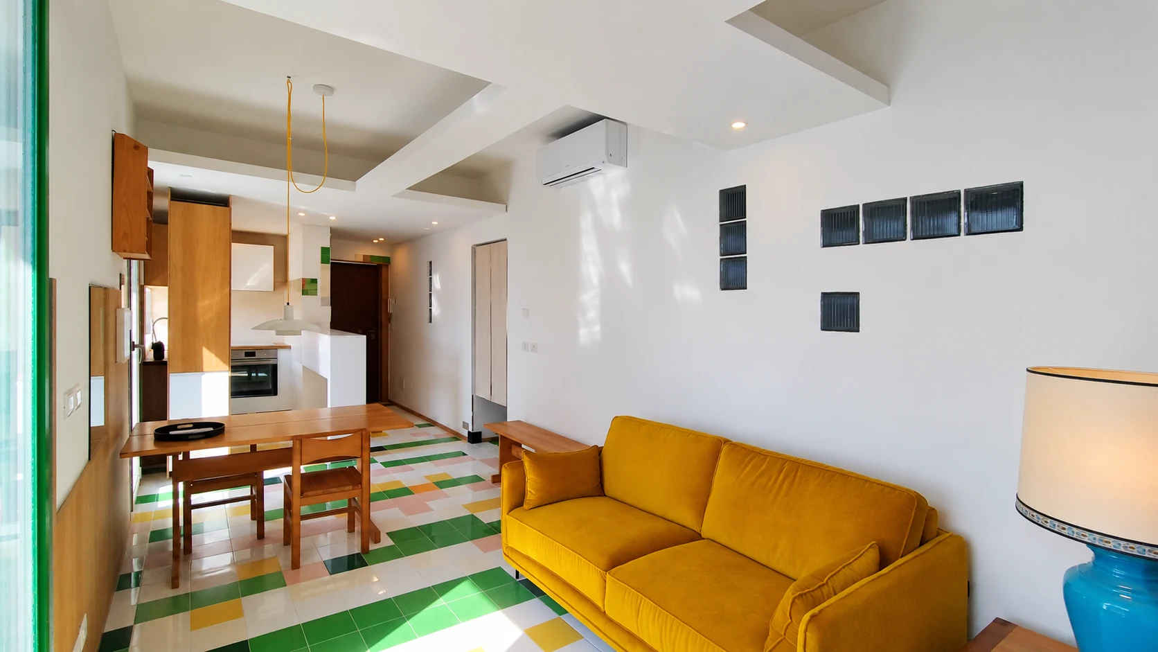 Soggiorno con divano giallo, inserti in vetrocemento e geometrie del soffitto, animato dai riflessi della luce naturale.